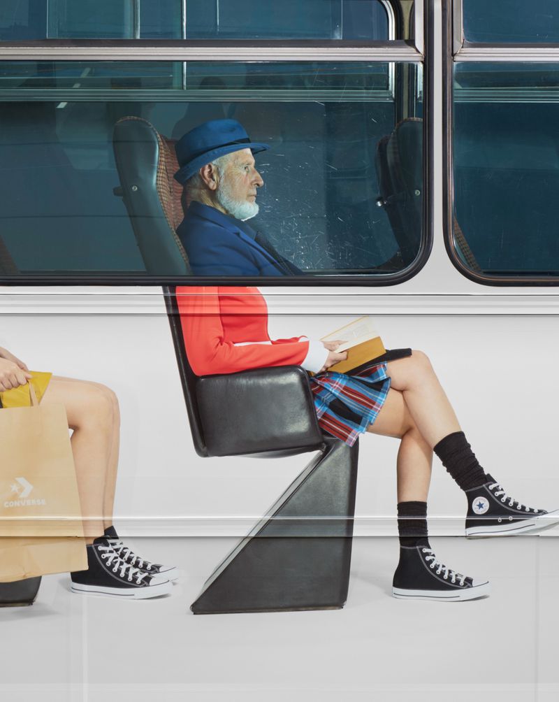 Elderly man in blue hat and suit jacket rides public transportation with young adult woman in skirt and sneakers; minimal background.