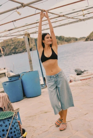 A young woman stands on a dock in a black bikini top and denim culottes, arms raised, in a sun-drenched Mediterranean seaside scene.