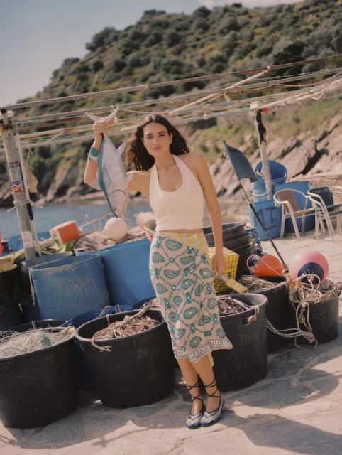 Young woman holds a large fish on a sunny pier, wearing a halter top, paisley skirt, and lace-up flats, with a rocky hillside in the background.