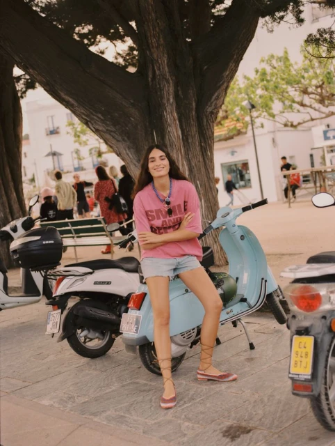 A young woman poses on a light blue vintage scooter with other scooters nearby, beneath a large tree in an outdoor public setting.