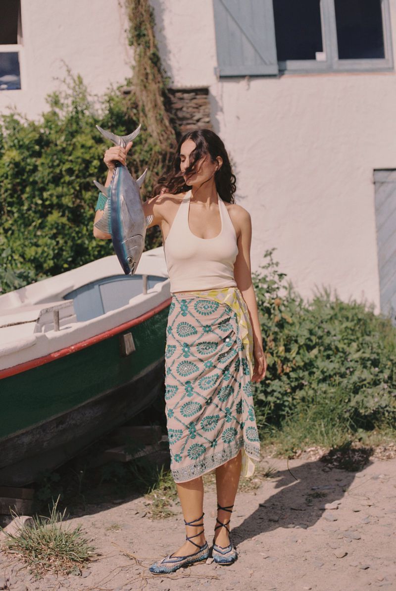 A woman in a paisley skirt holds a large tuna fish near a boat, with a whitewashed building and green foliage in the background.