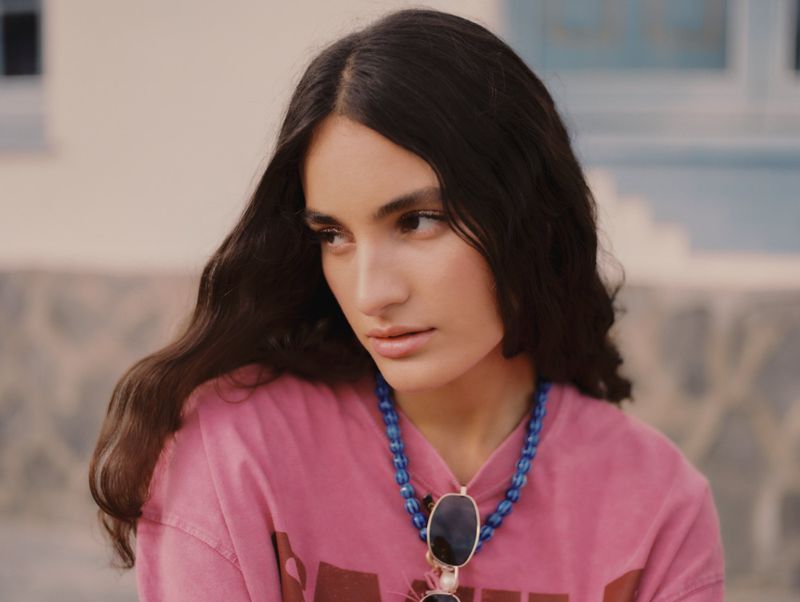 Model Marimar Cardenas posing in a pink graphic t-shirt and blue beaded necklace for Ulanka S/S 25.