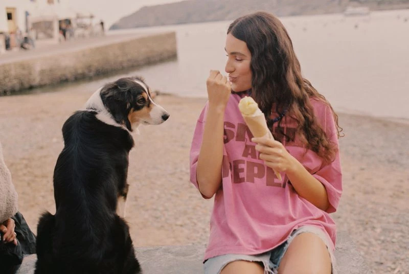 Woman enjoying ice cream with her dog by the ocean on a sunny day. A pink oversized t-shirt and denim shorts complete her relaxed look.