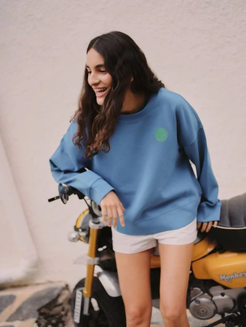 Smiling woman in a blue sweatshirt with a green circle design leaning on a yellow and black mini motorcycle in front of a wall.