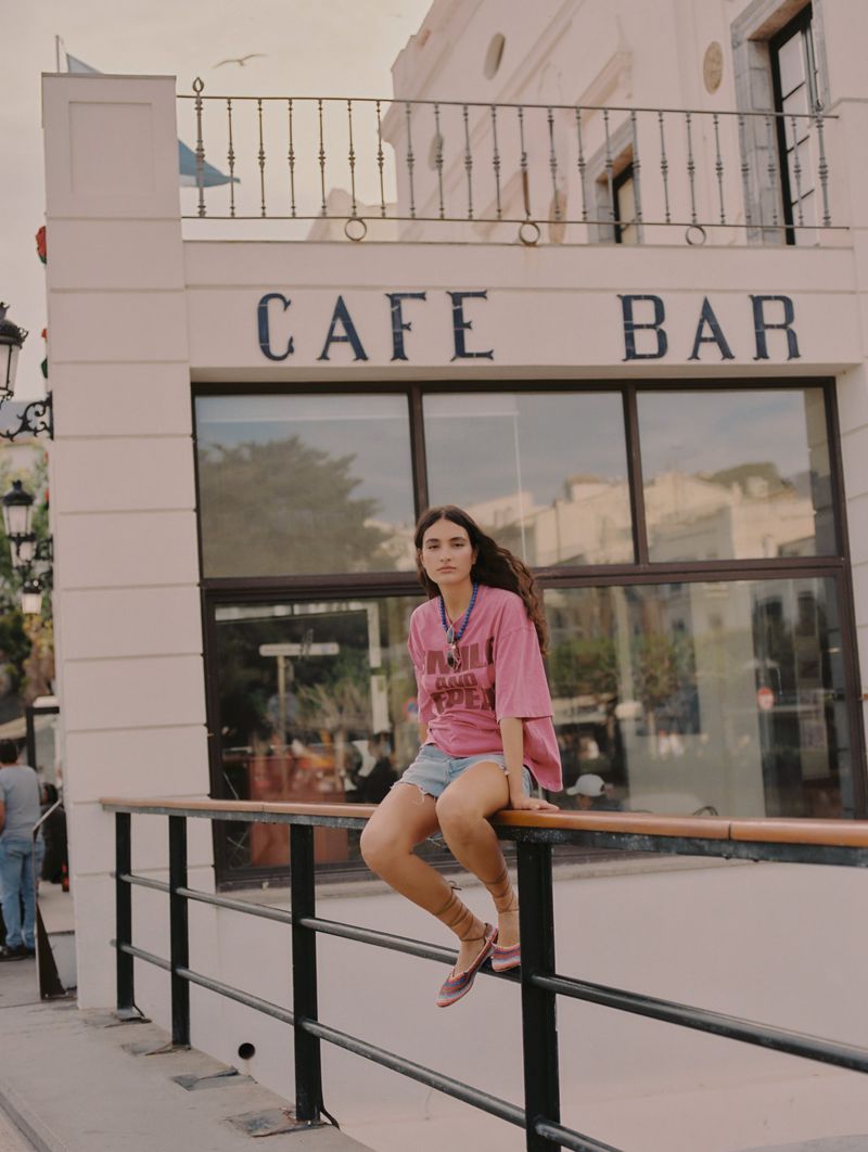 Young adult woman sits on a railing in front of a cafe bar. She wears a pink t-shirt and denim shorts. The building is white.