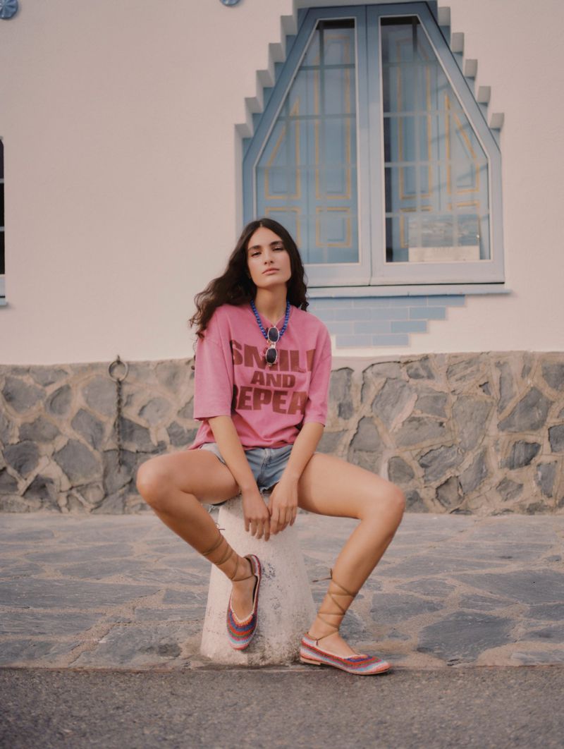 A woman in a pink t-shirt and blue jean shorts sits on a stone pillar outdoors with a white building and window in the background.