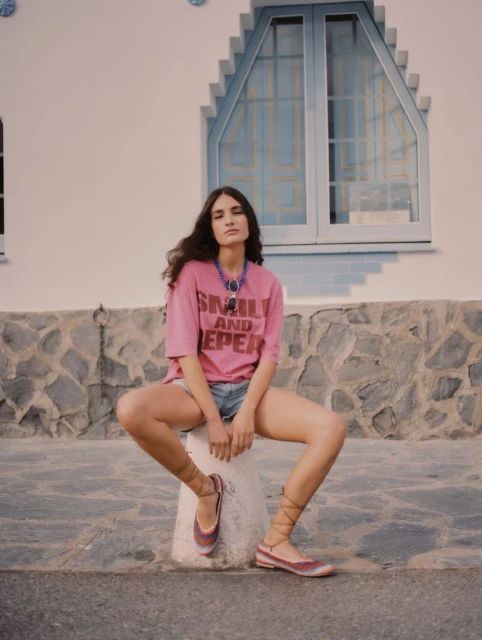 A woman in a pink t-shirt and blue jean shorts sits on a stone pillar outdoors with a white building and window in the background.