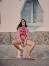 Marimar Cardenas sits on a bollard wearing a pink graphic t-shirt, denim shorts, and lace-up flats in front of a white building.