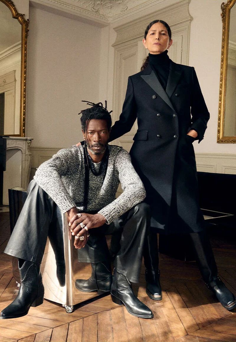 Two people pose indoors: a Black man in sweater and leather pants seated on a chrome cube; a woman in a black coat stands beside him.