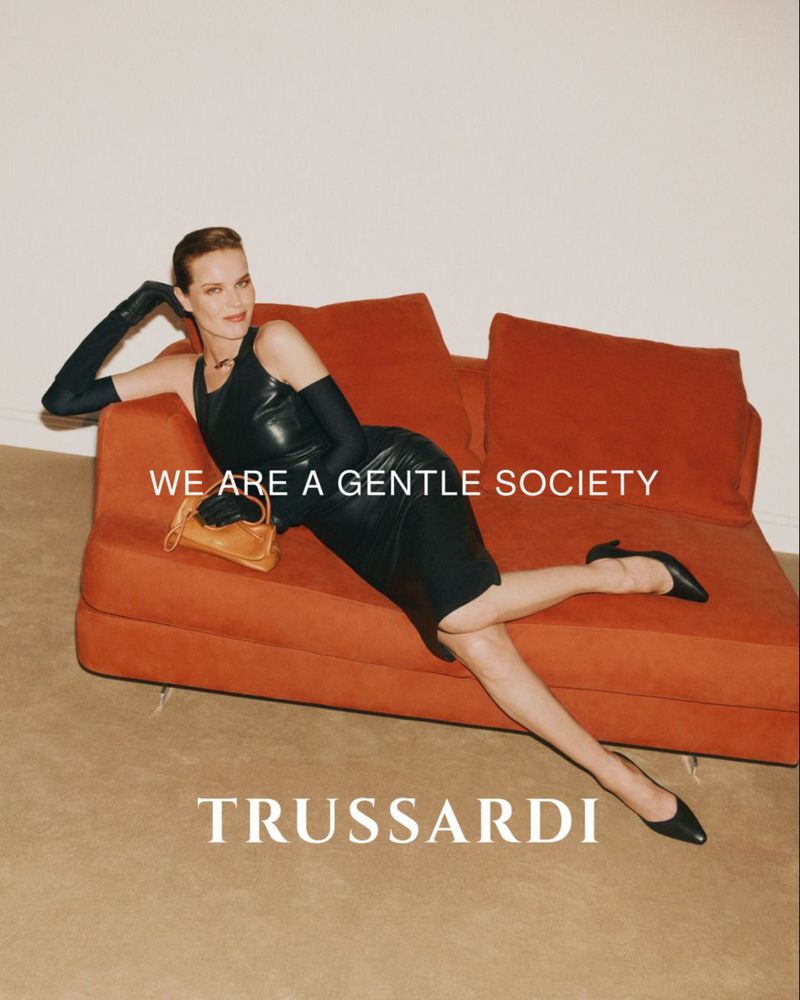 A woman in a black dress and gloves reclines on a rust-colored couch with a handbag, showcasing a fashion-forward and relaxed pose.