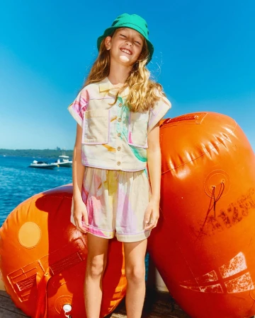 Smiling young girl outdoors in a colorful shirt and shorts with a green bucket hat, posing near large orange buoys on a sunny day.