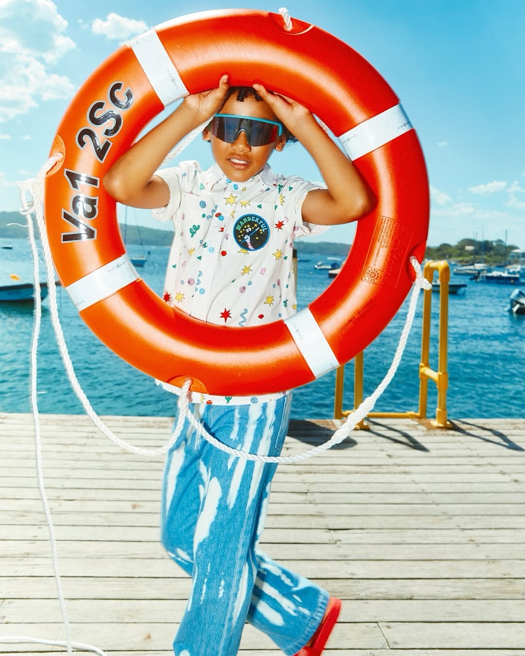 Child in patterned shirt and blue pants poses playfully with an orange life preserver in a sunny coastal setting near boats and the ocean.