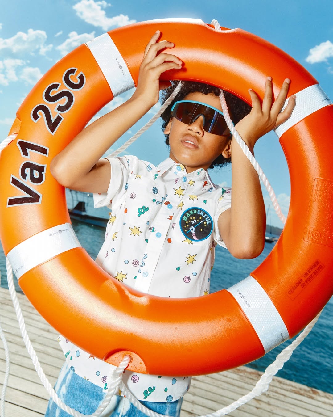 Young boy with sunglasses and a patterned shirt playfully holds an orange life preserver on a sunny dockside setting.