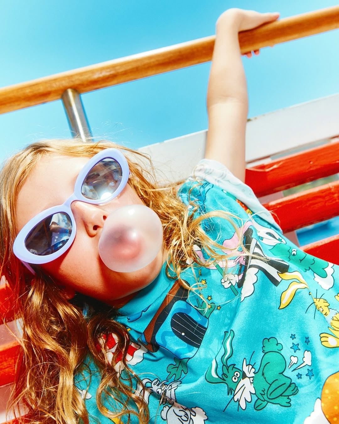 Child with bubble gum lying on a bench wearing sunglasses and a blue patterned shirt against a blue sky in bright daylight.