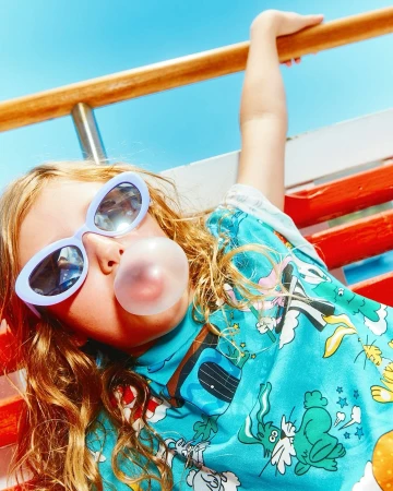 Child with bubble gum lying on a bench wearing sunglasses and a blue patterned shirt against a blue sky in bright daylight.