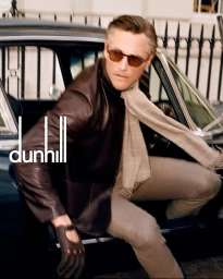 Man wearing a brown leather jacket, sunglasses, and driving glove stepping out of a vintage convertible for the Dunhill campaign.