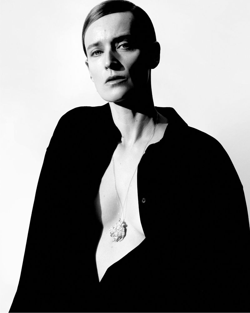 Monochromatic portrait of a model with slicked-back hair, wearing a black top and a silver necklace in a bright studio setting.