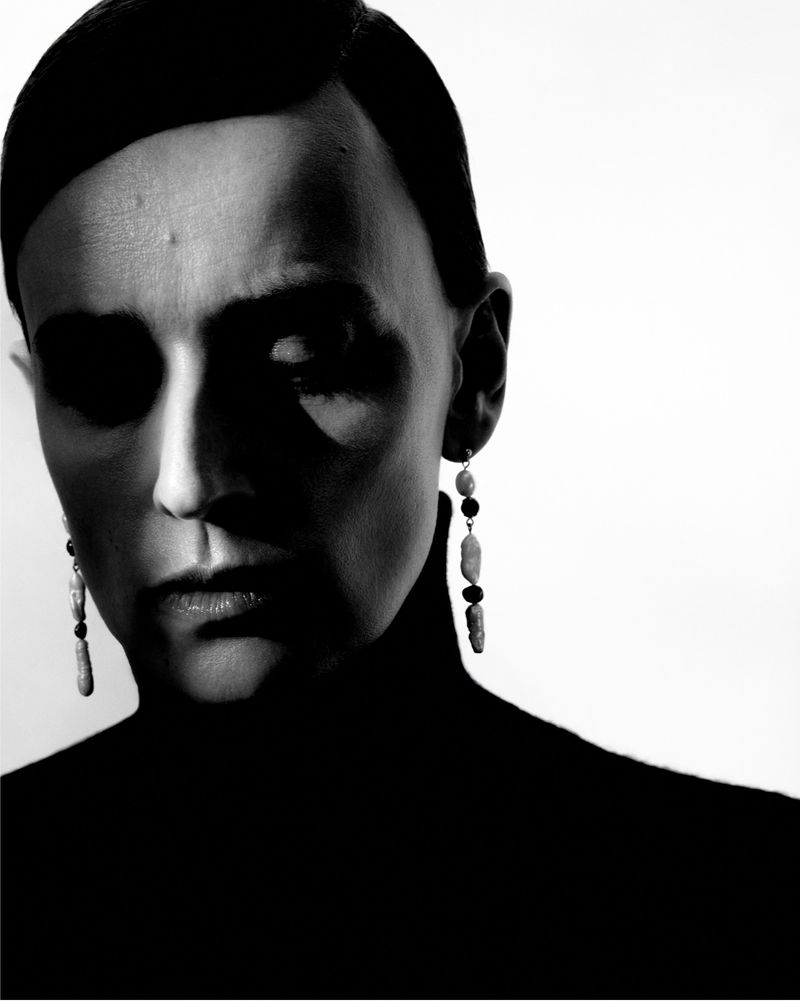 Monochrome portrait of adult female with short dark hair, wearing a black turtleneck, featuring dangling bead earrings against white.