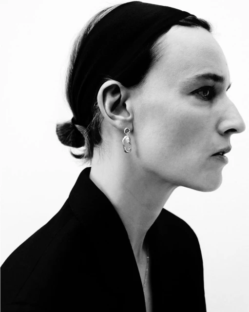 Monochromatic portrait of an adult female in profile wearing a black headband, jacket, and statement earrings against a white backdrop.