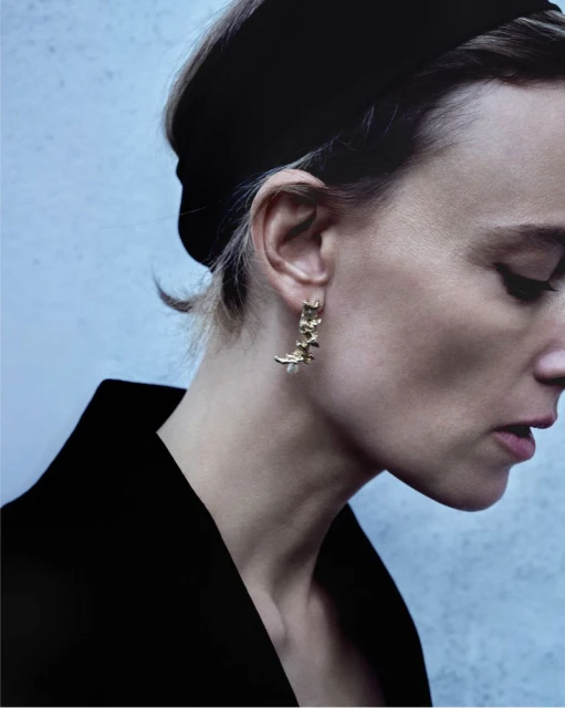 Portrait of a stylish adult female model showcasing gold floral-shaped earrings with a pearl, wearing a black dress and headband.