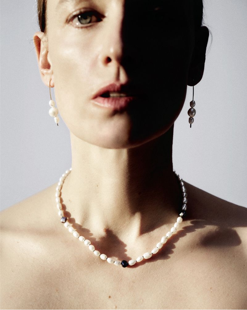 Portrait of a fair-skinned woman with pearl jewelry including earrings and necklace, shadows across face, neutral background.