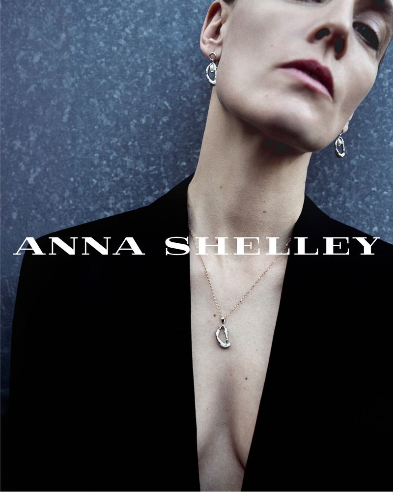 Anne-Catherine Lacroix in a black deep-V blazer wearing a sculpted silver pendant necklace and drop earrings against a slate wall.