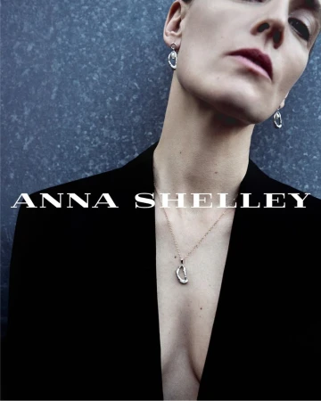 Anne-Catherine Lacroix in a black deep-V blazer wearing a sculpted silver pendant necklace and drop earrings against a slate wall.