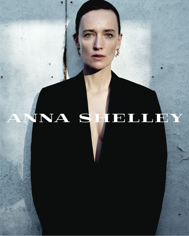 Portrait of a fair-skinned woman with green eyes wearing a dark blazer and earrings against a muted blue-gray background, fashion style.