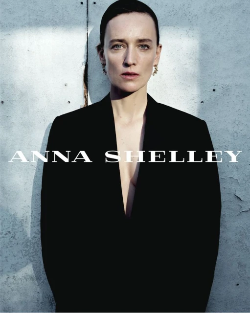 Portrait of a fair-skinned woman with green eyes wearing a dark blazer and earrings against a muted blue-gray background, fashion style.