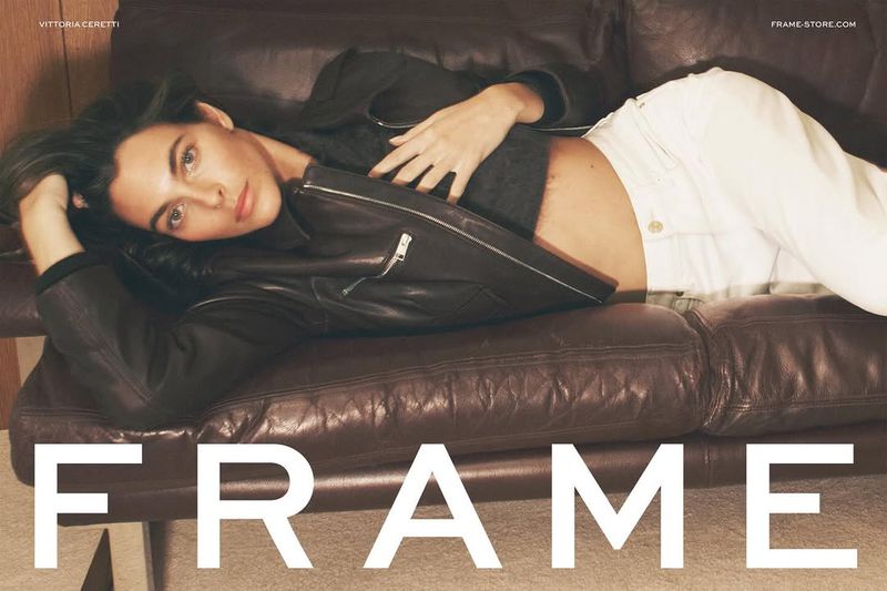 Brunette model Vittoria Ceretti lounges on a brown leather couch wearing a black jacket and white jeans, promoting FRAME brand fashion.