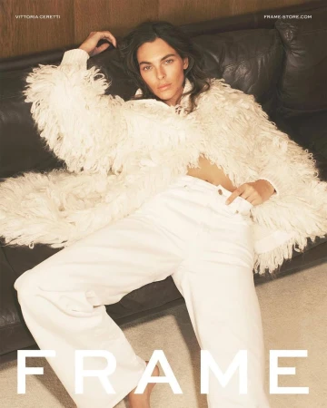 Brunette woman in a shaggy white jacket and white denim pants reclines on a dark leather couch, photographed indoors in soft lighting.
