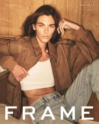 Model Vittoria Ceretti wearing an oversized textured brown jacket, white crop top, and blue jeans against a wood-paneled wall.