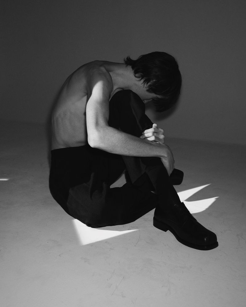 Shirtless model sitting curled up on the studio floor wearing dark trousers and black dress shoes under sharp geometric shadows.