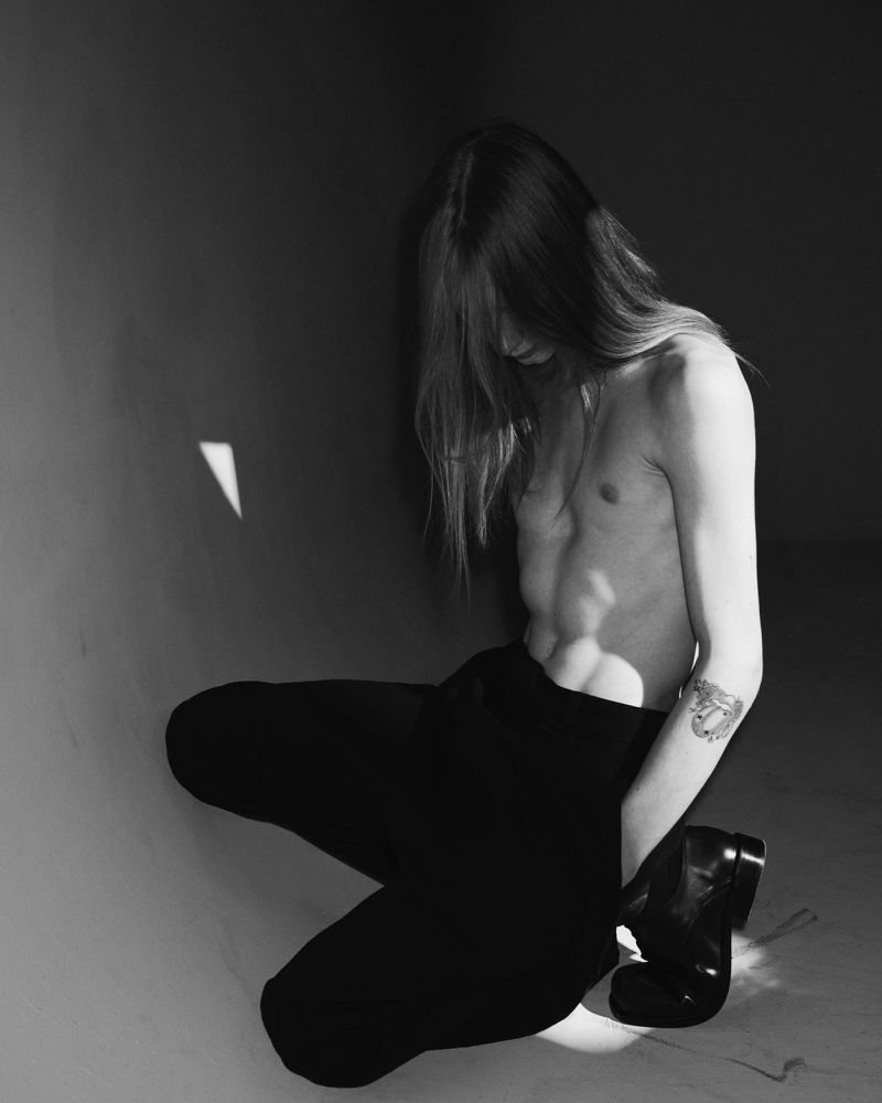 Black and white portrait of a shirtless young adult kneeling in black pants and boots with long hair near a wall with light.