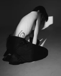 Black and white photo of a topless model kneeling in tailored black trousers, displaying a calligraphic tattoo on their side.
