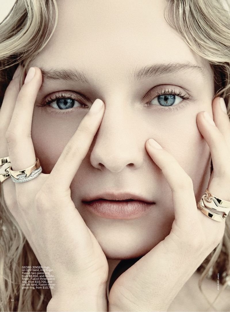 Close-up of a fair-skinned adult woman with blonde wavy hair and blue eyes, wearing gold rings and holding her hands near her face.