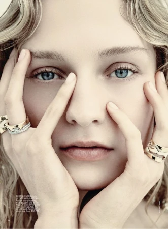 Close-up portrait of a woman framing her face with hands wearing statement gold and diamond rings against a neutral backdrop.