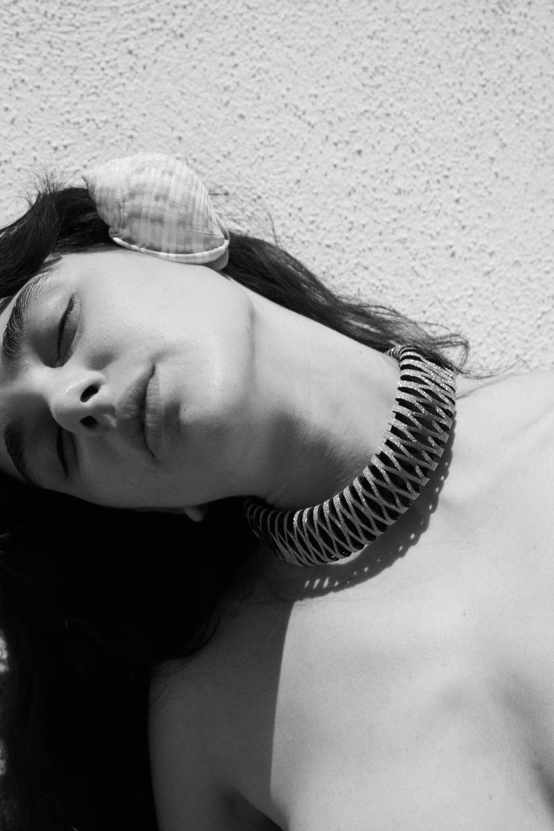 Black and white portrait of a woman lying in the sun with a large seashell on her head and a wide, coiled metal necklace.