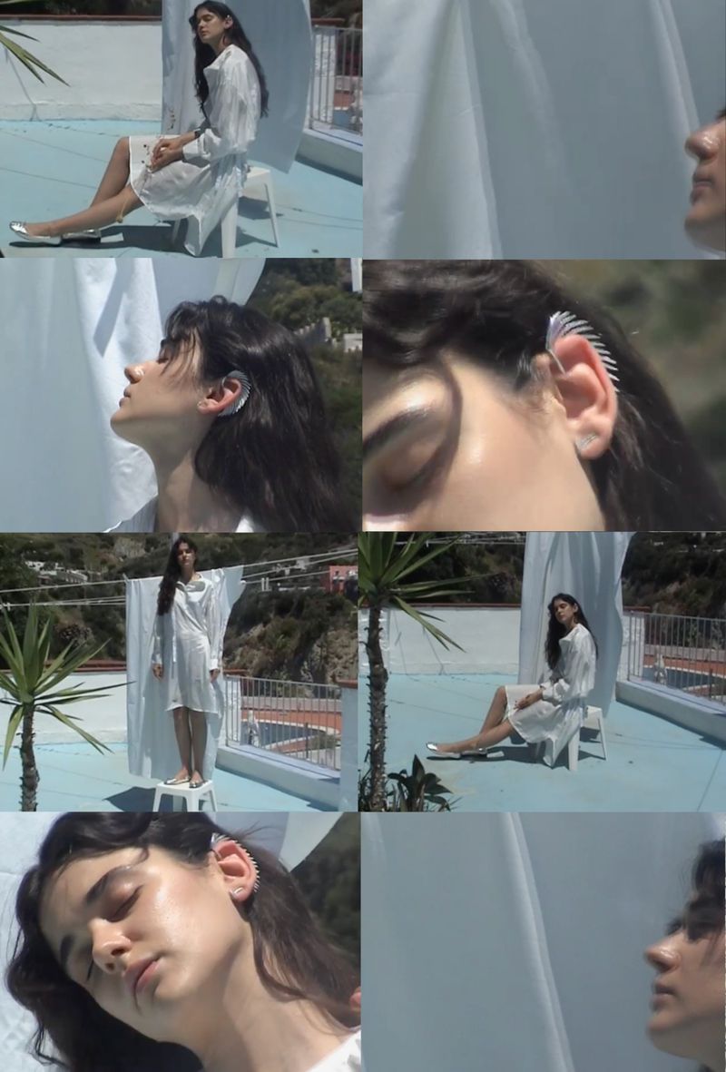 A woman in a white dress is on a sunny rooftop, leaning on a white rail, wearing statement ear jewellery, and sheer white fabric as backdrop.