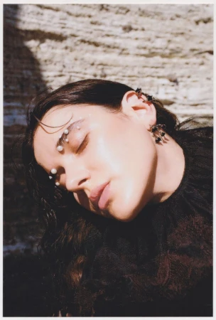 Portrait of a young woman with pearl embellishments on her face, black earrings, leaning against a textured rock surface.