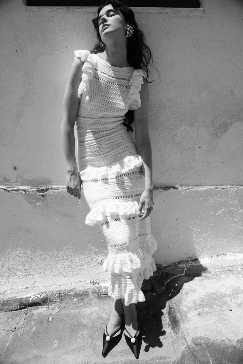 Monochrome fashion shot of a young woman in a white crochet dress leaning against a wall, complemented by black pointed-toe heels.