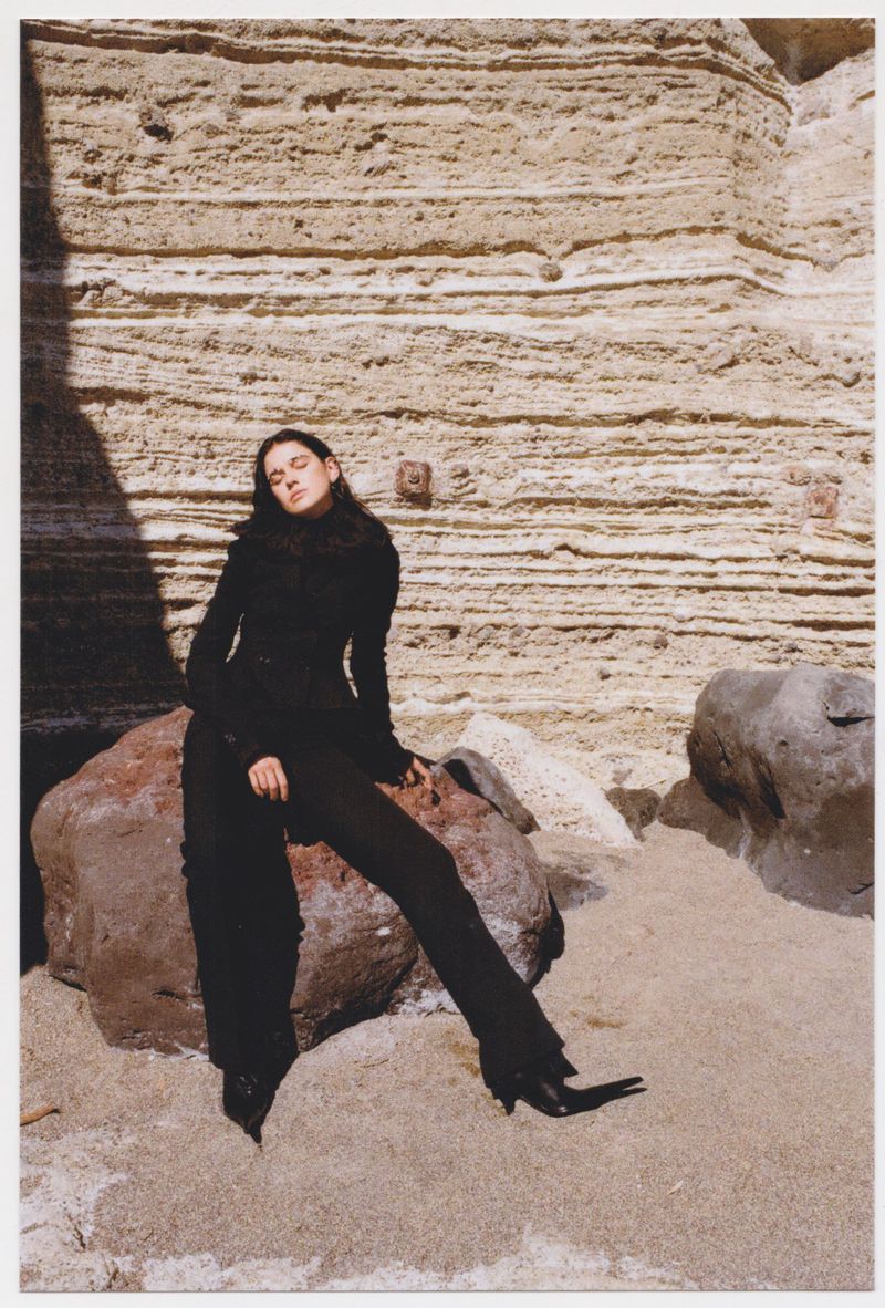 A fashion photo shows a young woman in a black outfit sitting on a rock on a sandy surface in front of a light brown rock formation.