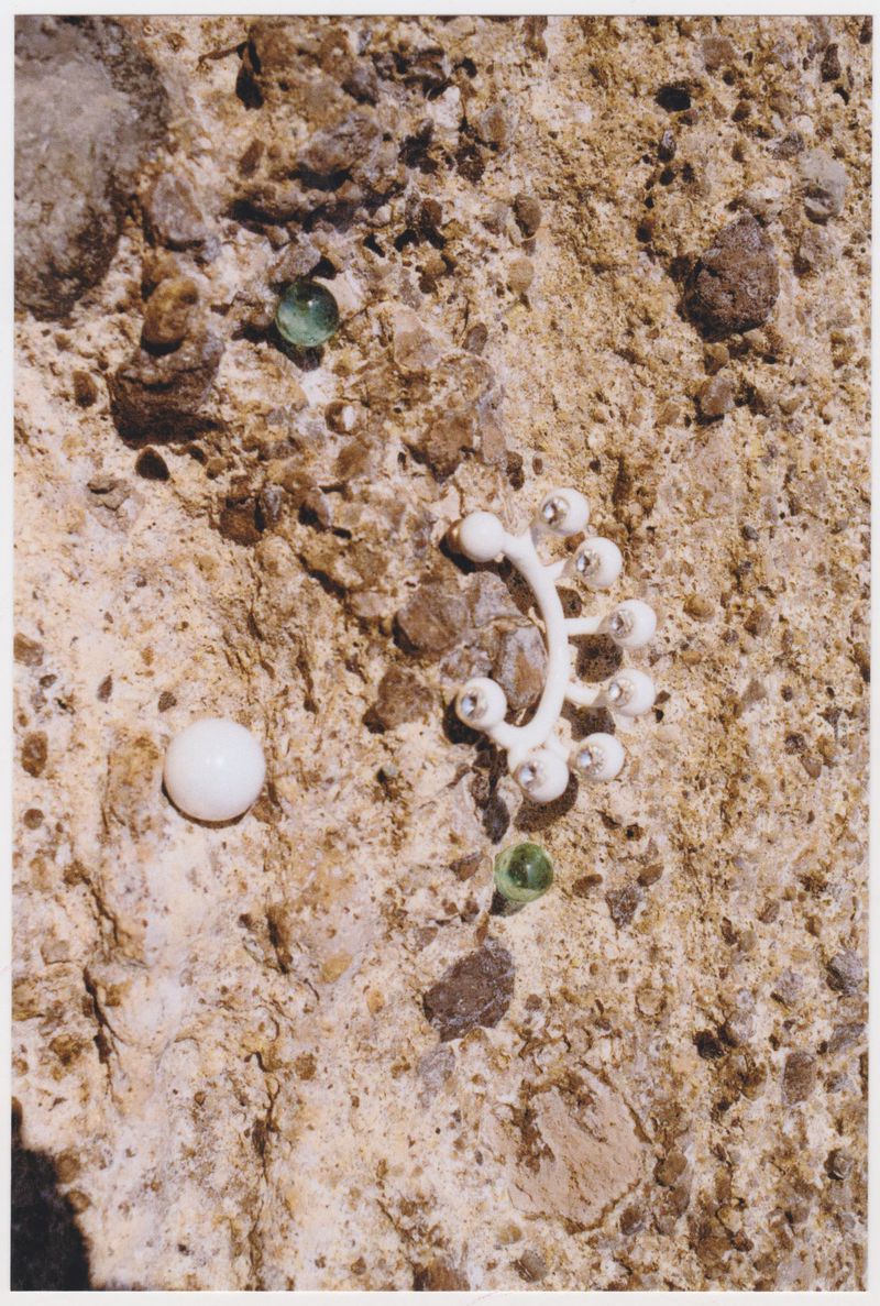 Overhead shot of white plastic joint model, green glass marbles, and small stones on a tan rock-like surface in natural light.