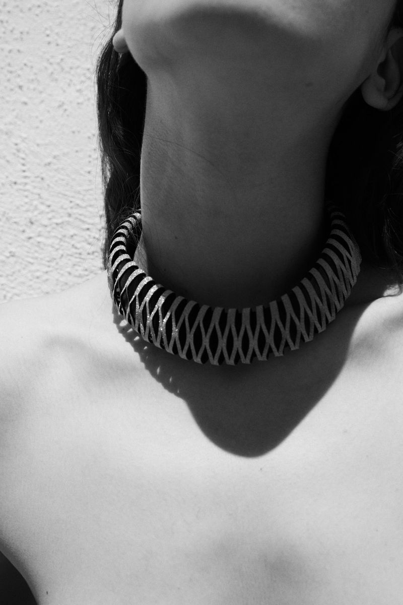 Black and white studio portrait of a woman's neck and shoulders wearing a sculptural statement necklace with shadow on the wall.