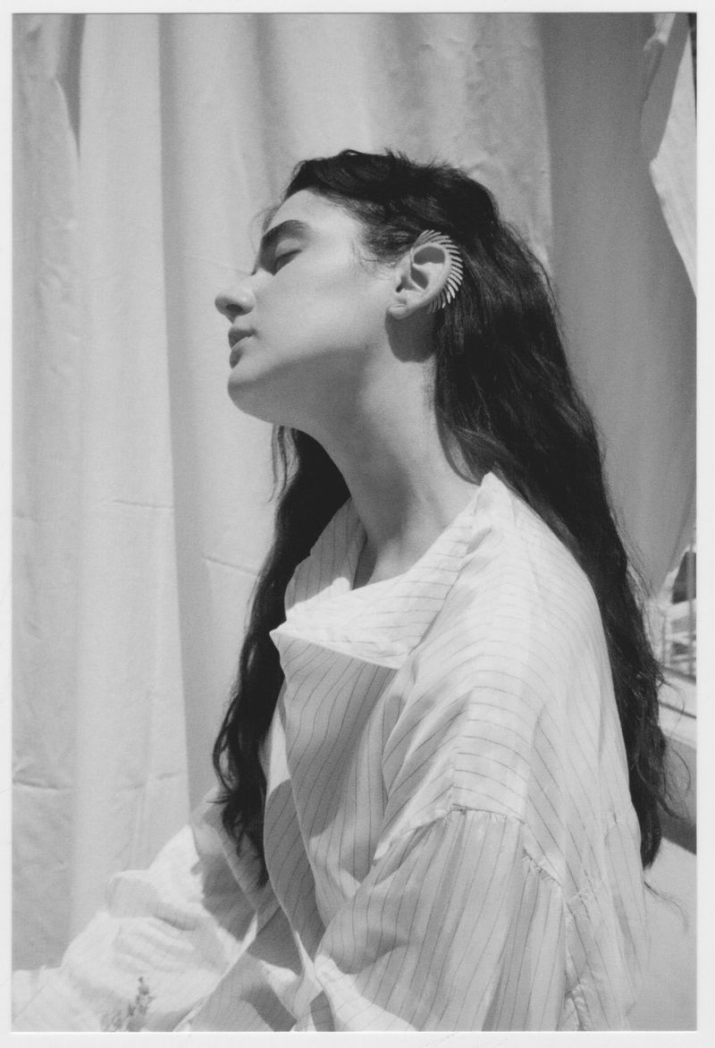 Portrait of a woman in a pinstripe shirt with an ear cuff, bathed in natural light against a draped white fabric backdrop.