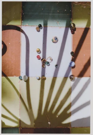 Glass marbles and a jewelry ring scattered across square pastel tiles under a sharp palm frond shadow.