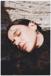 Close-up portrait of a woman with pearls on her eyebrows wearing a dark ruffled top against a textured stone wall.