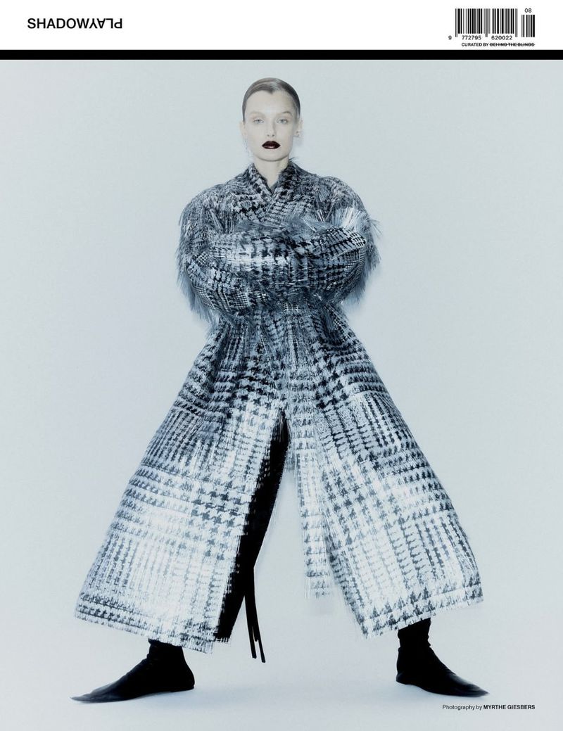 Fashion editorial shot featuring a woman wearing a black and white patterned coat and black boots against a light background in a studio.