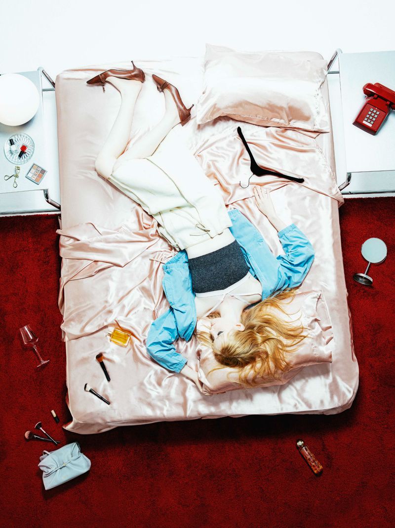 Overhead studio shot of a woman reclining on a bed with makeup, shoes, and accessories in disarray, creating a relaxed, artistic scene.