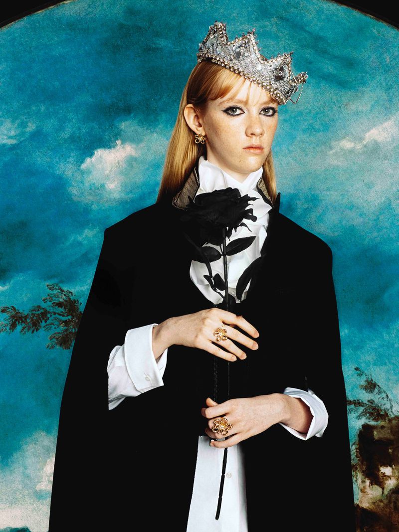Portrait of a woman in a cape and crown, holding a black rose against a painted backdrop in a fashion or editorial photograph.
